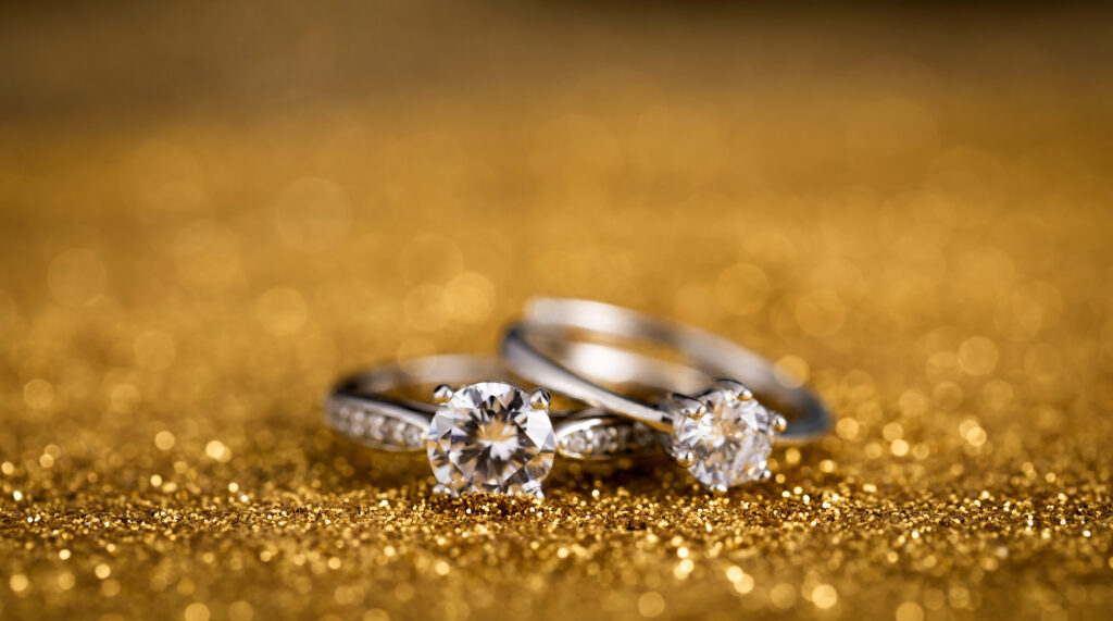 Close-up of two round brilliant-cut moissanite engagement rings with silver bands, placed on a sparkling golden surface, highlighting their radiant clarity and elegance.