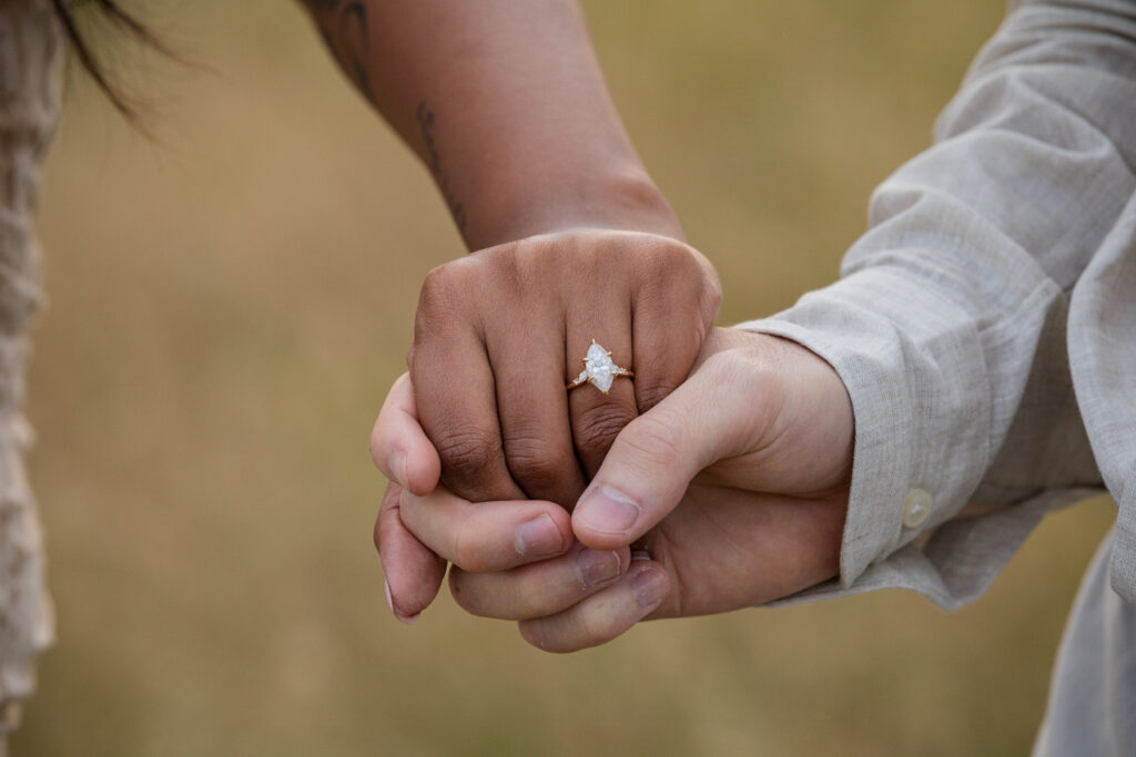 Close-up of an engaged couple holding hands, featuring a marquise-cut moissanite engagement ring with a gold band on the woman's hand, set against a blurred natural background.