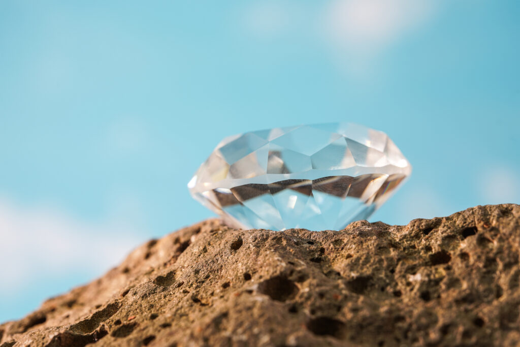 Close-up of a faceted moissanite gemstone resting on a textured rock surface with a bright blue sky background, showcasing its clarity and brilliance.