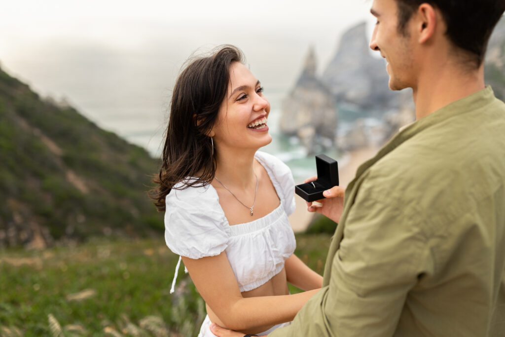 Man proposing with a moissanite engagement ring in a black box to a joyful woman on a scenic cliffside overlooking the ocean, capturing a romantic outdoor moment.