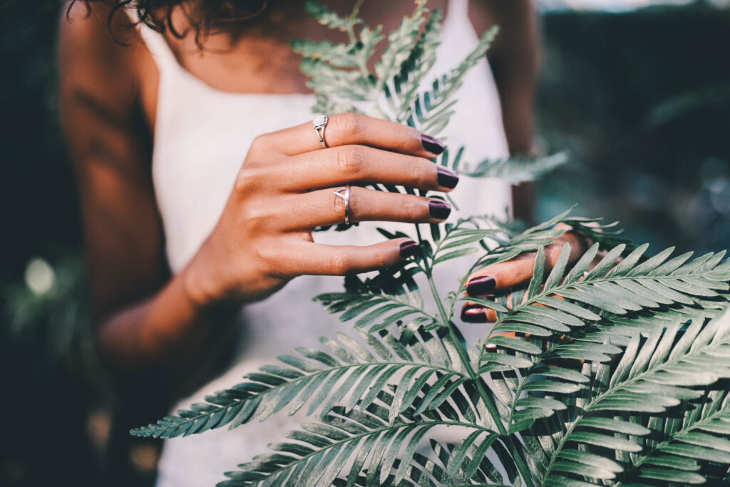 Close-up of a woman's hand adorned with minimalist moissanite rings, gently touching green fern leaves, symbolizing nature-inspired sustainable jewelry.