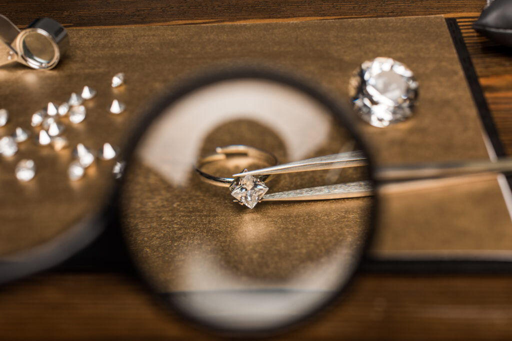 Close-up view of a jeweler examining a princess-cut moissanite engagement ring with tweezers through a magnifying glass, highlighting precision craftsmanship and gemstone quality.