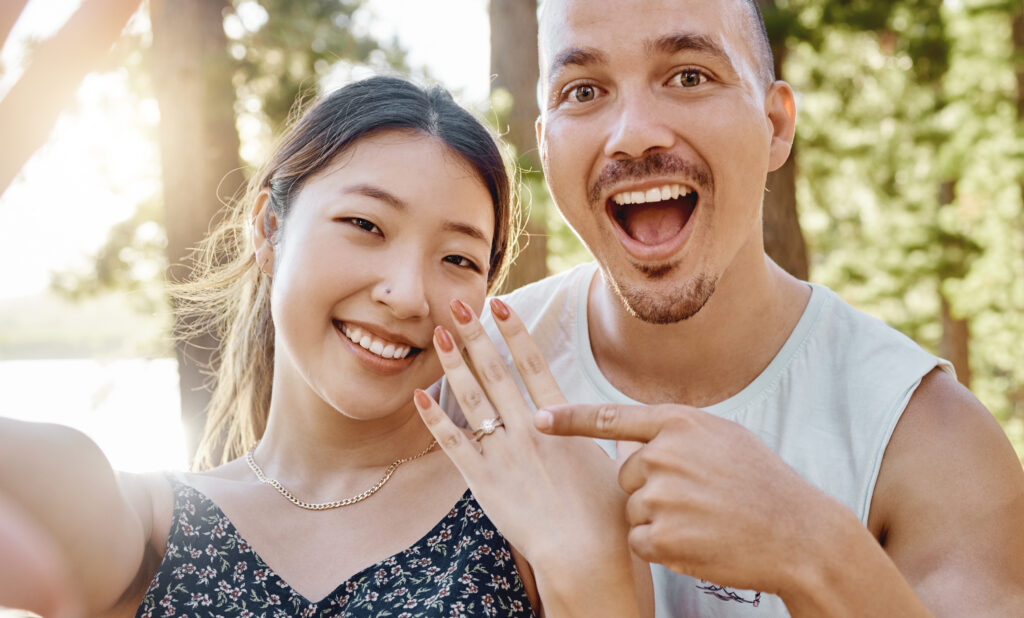 Happy engaged couple taking a selfie outdoors, with the woman proudly displaying her moissanite engagement ring, capturing the joy of their special moment.