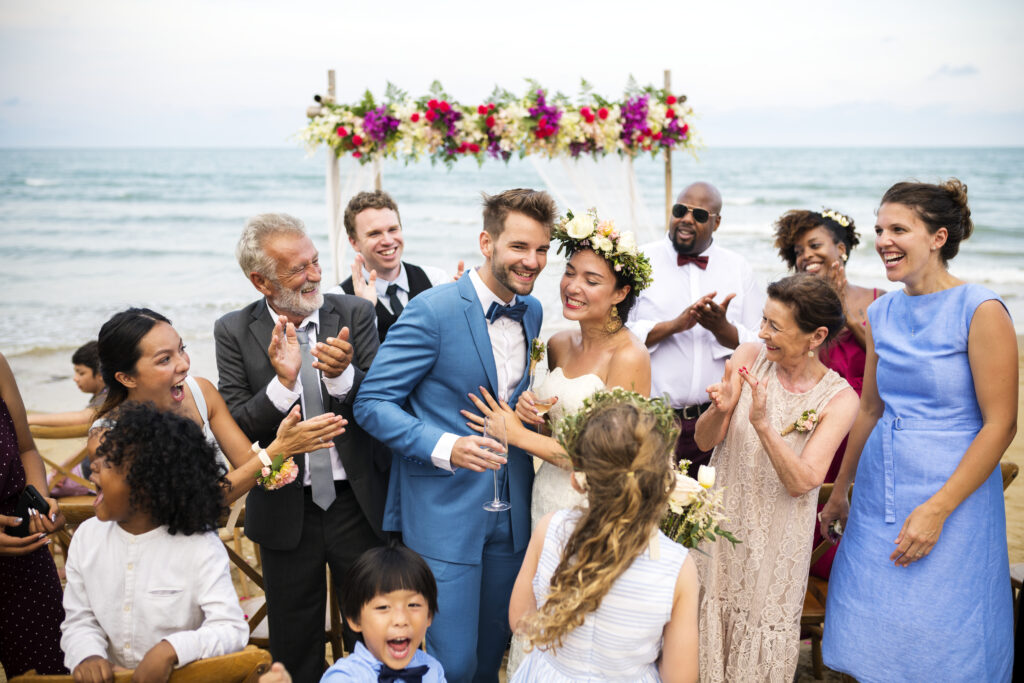 Joyful newlywed couple celebrating their beach wedding surrounded by family and friends, with the bride wearing a floral crown and a moissanite engagement ring.